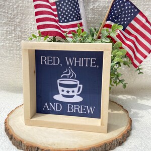 Pu&ograve; includere: Un cartello di legno con sfondo blu e testo bianco che recita "Red, White, and Brew". Il cartello presenta una tazza di caff&egrave; bianca con stelle rosse, bianche e blu. Il cartello &egrave; appoggiato su una fetta di legno con due piccole bandiere americane su bastoni dietro di esso.