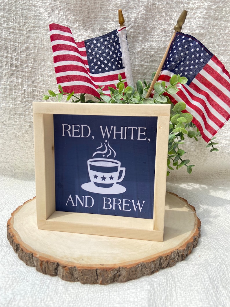 Pu&ograve; includere: Un cartello di legno con sfondo blu e testo bianco che recita "Red, White, and Brew". Il cartello presenta una tazza di caff&egrave; bianca con stelle rosse, bianche e blu. Il cartello &egrave; appoggiato su una fetta di legno con due piccole bandiere americane su bastoni dietro di esso.