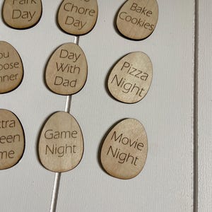 Op de afbeelding: Houten eivormige tokens met verschillende gezinsactiviteiten erop geschreven, zoals "Parkdag", "Geen klusjesdag", "Koekjes bakken", "Dag met papa", "Pizza avond", "Spelletjesavond" en "Filmavond".