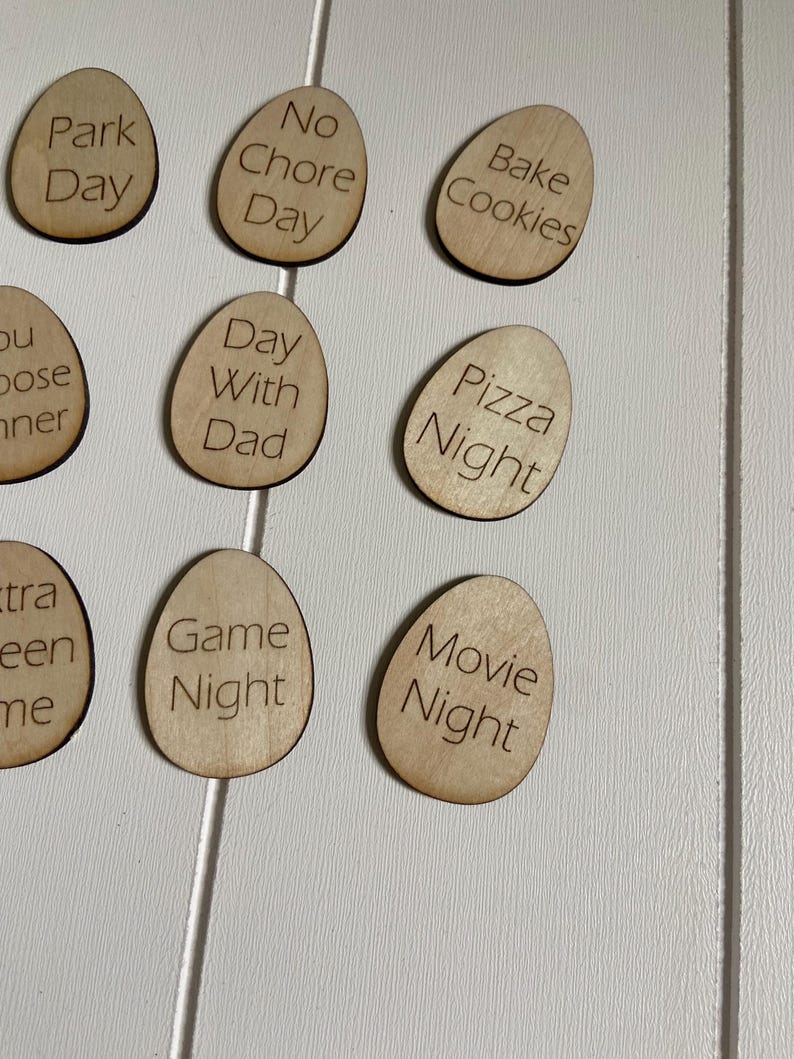 Op de afbeelding: Houten eivormige tokens met verschillende gezinsactiviteiten erop geschreven, zoals "Parkdag", "Geen klusjesdag", "Koekjes bakken", "Dag met papa", "Pizza avond", "Spelletjesavond" en "Filmavond".