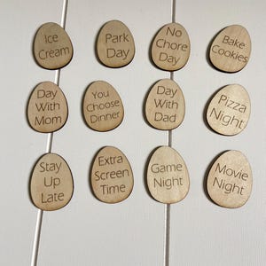 Op de afbeelding: Twaalf houten eieren met verschillende activiteiten erop geschreven, zoals "IJs", "Dagje uit", "Geen klusjes", "Koekjes bakken", "Dag met mama", "Jij kiest het diner", "Dag met papa", "Pizza avond", "Laat opblijven", "Extra schermtijd", "Spelletjesavond" en "Filmavond".