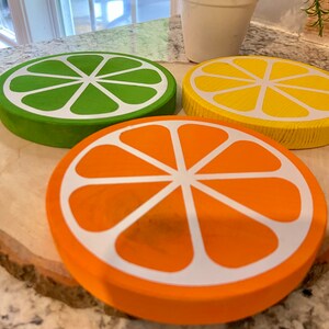 Puede incluir: Tres posavasos de madera con forma de rodajas de c&iacute;tricos. Los posavasos son verdes, amarillos y naranjas con dise&ntilde;os pintados en blanco. Los posavasos est&aacute;n sobre una superficie de madera.