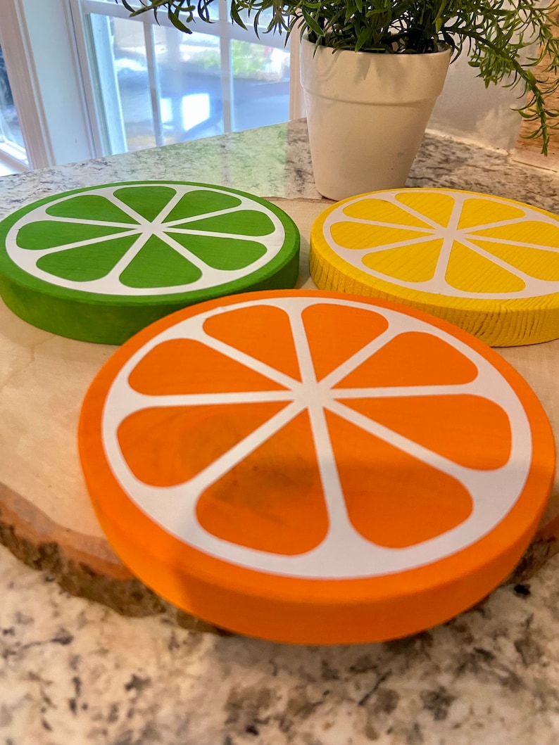 Puede incluir: Tres posavasos de madera con forma de rodajas de c&iacute;tricos. Los posavasos son verdes, amarillos y naranjas con dise&ntilde;os pintados en blanco. Los posavasos est&aacute;n sobre una superficie de madera.