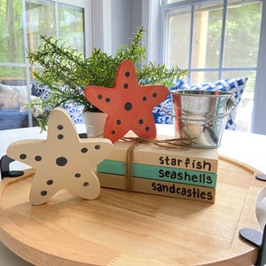 May include: A wooden tray with three stacked books. The books are painted with the words "starfish", "seashells", and "sandcastles". There are two starfish decorations on the tray, one orange and one beige.