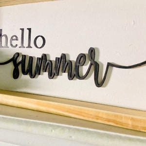 Op de afbeelding: Een wit houten bord met een zwarte houten uitsnijding van de woorden "hello summer".