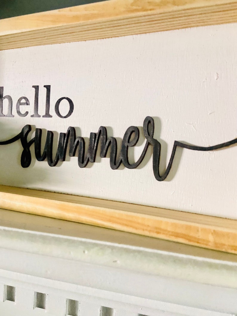 Op de afbeelding: Een wit houten bord met een zwarte houten uitsnijding van de woorden "hello summer".