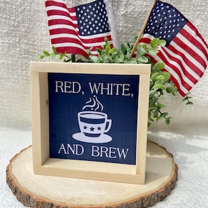 Pu&ograve; includere: Un cartello di legno con sfondo blu e testo bianco che recita "Red, White, and Brew". Una tazza di caff&egrave; bianca con stelle rosse, bianche e blu &egrave; raffigurata sul cartello. Il cartello &egrave; appoggiato su una fetta di legno.
