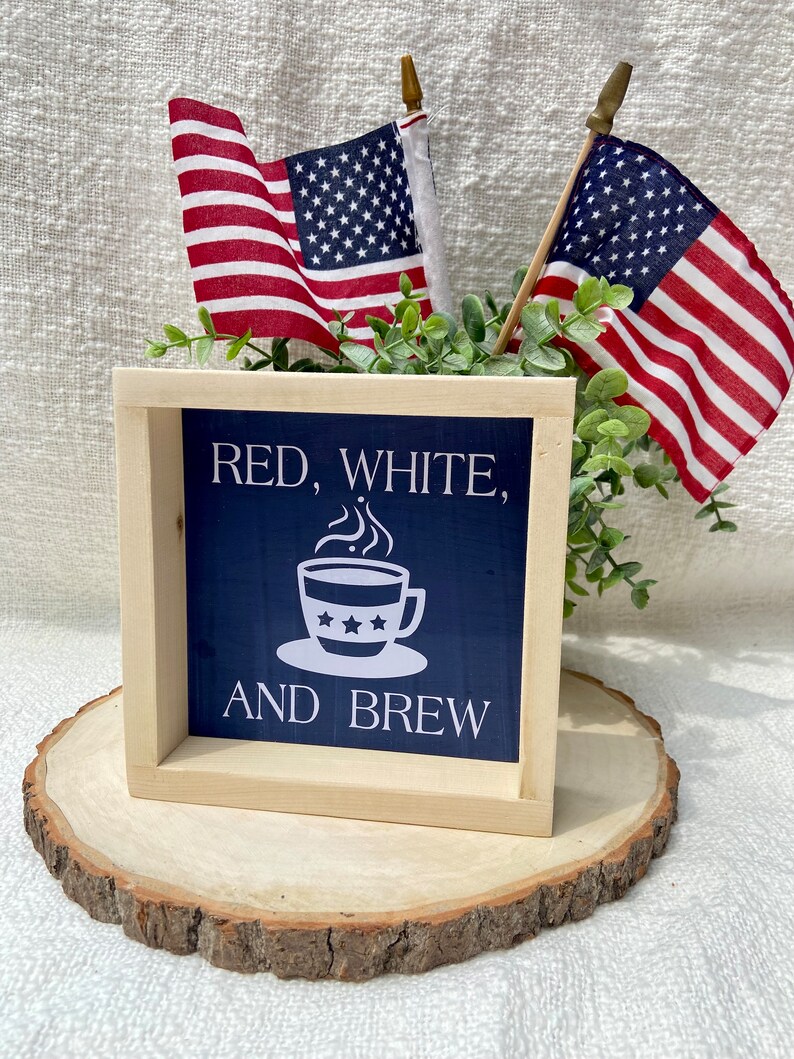 Pu&ograve; includere: Un cartello di legno con sfondo blu e testo bianco che recita "Red, White, and Brew". Una tazza di caff&egrave; bianca con stelle rosse, bianche e blu &egrave; raffigurata sul cartello. Il cartello &egrave; appoggiato su una fetta di legno.
