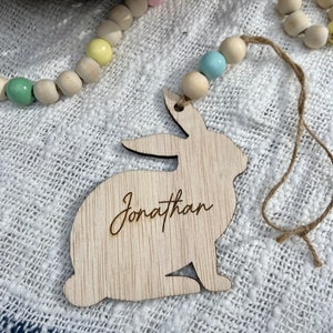 K&ouml;nnte beinhalten: Ausgeschnittener Holzhase mit dem eingravierten Namen Jonathan. Der Hase h&auml;ngt an einer Schnur aus Bindfaden mit pastellfarbenen Holzperlen.
