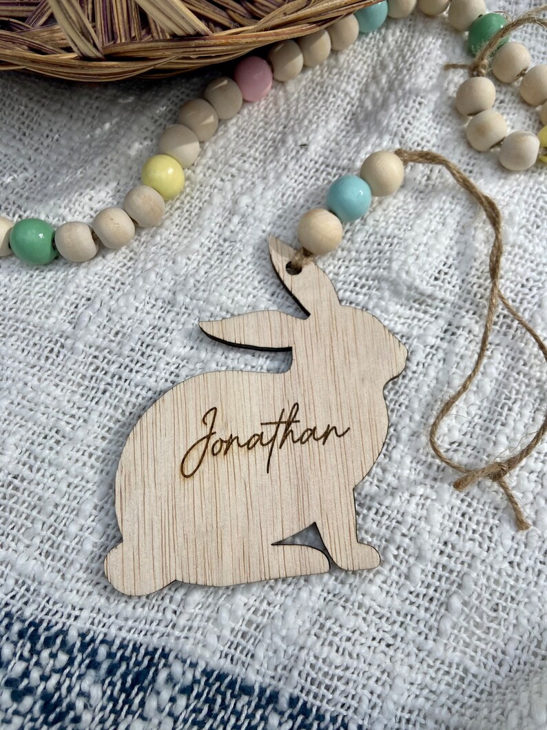 K&ouml;nnte beinhalten: Ausgeschnittener Holzhase mit dem eingravierten Namen Jonathan. Der Hase h&auml;ngt an einer Schnur aus Bindfaden mit pastellfarbenen Holzperlen.