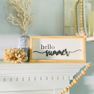 Op de afbeelding: Een houten bord met de tekst "hello summer" in zwarte cursieve letters. Het bord staat op een witte schouw met een witte koraal en een blauw-witte vaas met witte takken.