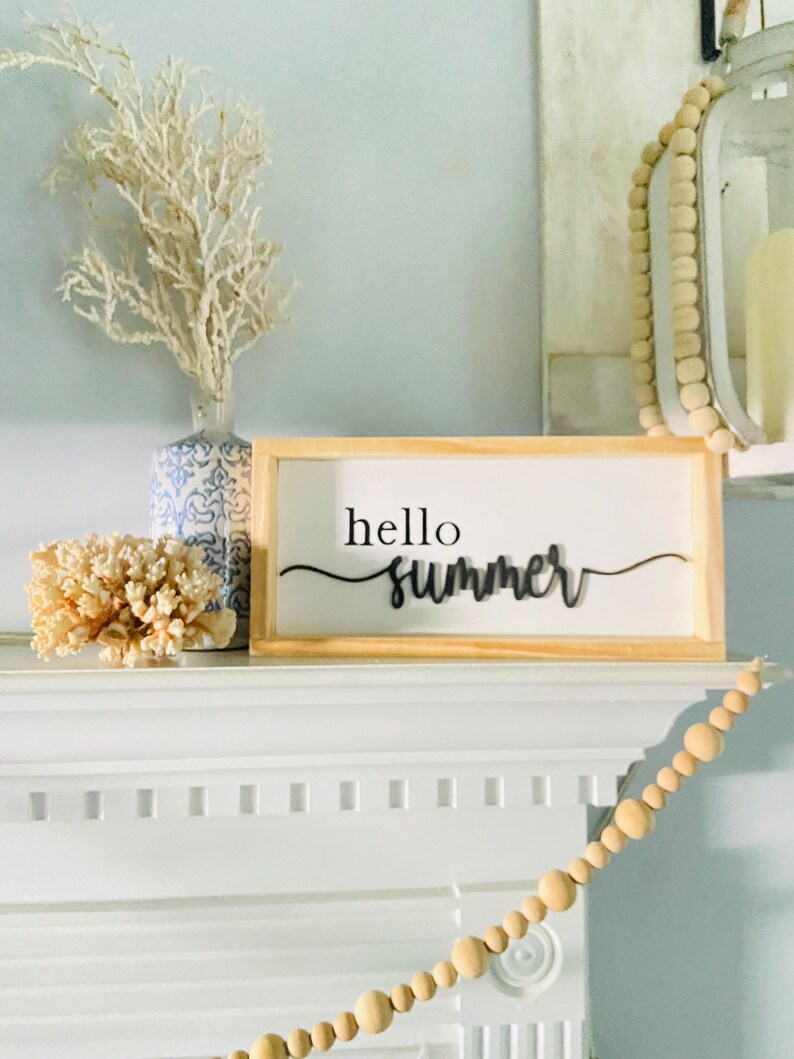 Op de afbeelding: Een houten bord met de tekst "hello summer" in zwarte cursieve letters. Het bord staat op een witte schouw met een witte koraal en een blauw-witte vaas met witte takken.