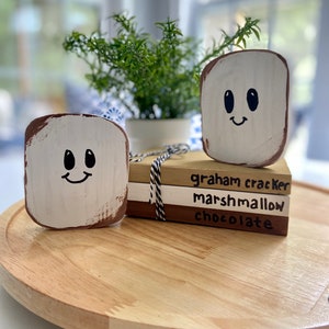 May include: A wooden craft project depicting a s'more. The craft is made of three blocks stacked on top of each other. The top block is white and has a smiley face drawn on it. The middle block is brown and says "marshmallow". The bottom block is tan and says "graham cracker".