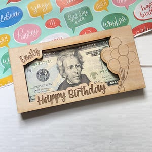 May include: A wooden money holder engraved with "Happy Birthday" and "Emily". It features a cutout to display a twenty-dollar note. A laser-cut design of balloons is on the right side of the holder.
