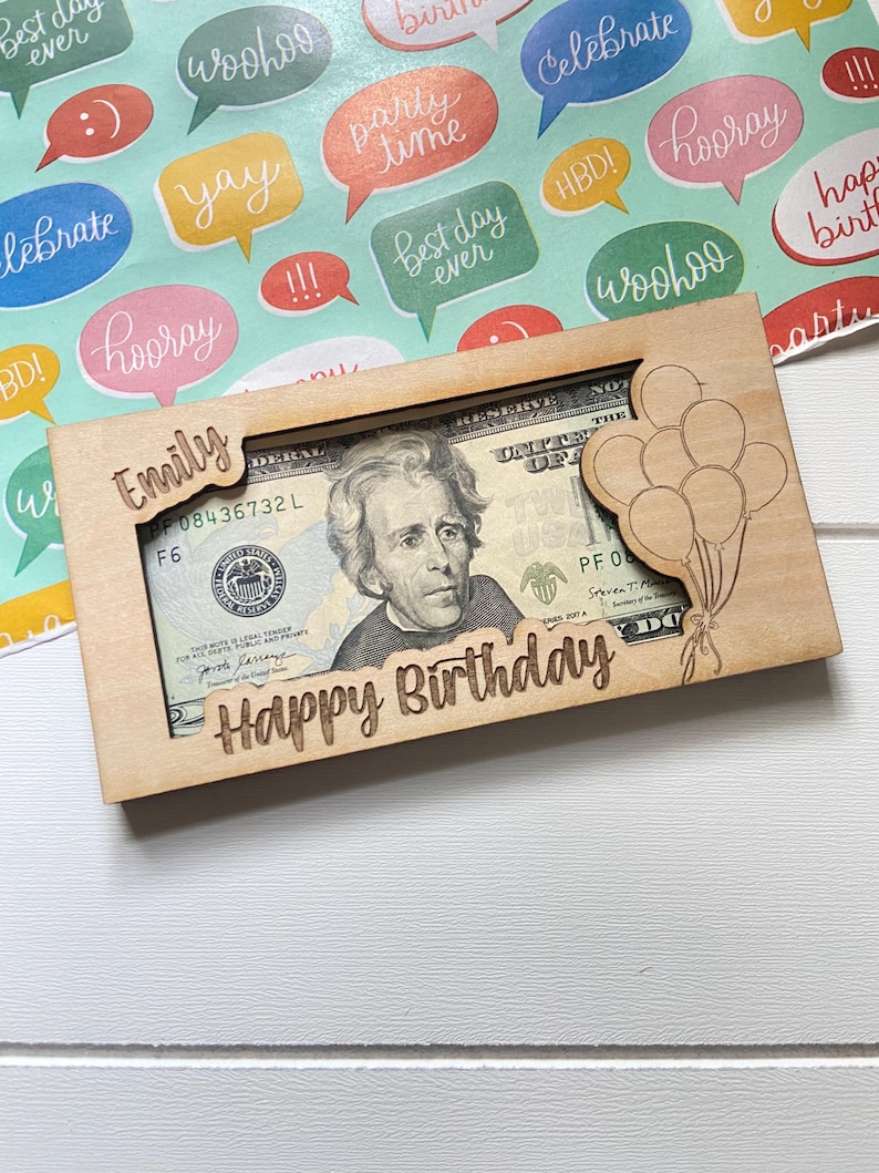May include: A wooden money holder engraved with "Happy Birthday" and "Emily". It features a cutout to display a twenty-dollar note. A laser-cut design of balloons is on the right side of the holder.