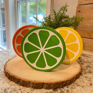 Puede incluir: Tres rodajas de madera pintadas para que parezcan frutas c&iacute;tricas. Las rodajas son naranja, verde y amarilla y est&aacute;n dispuestas sobre una rodaja de madera con corteza.