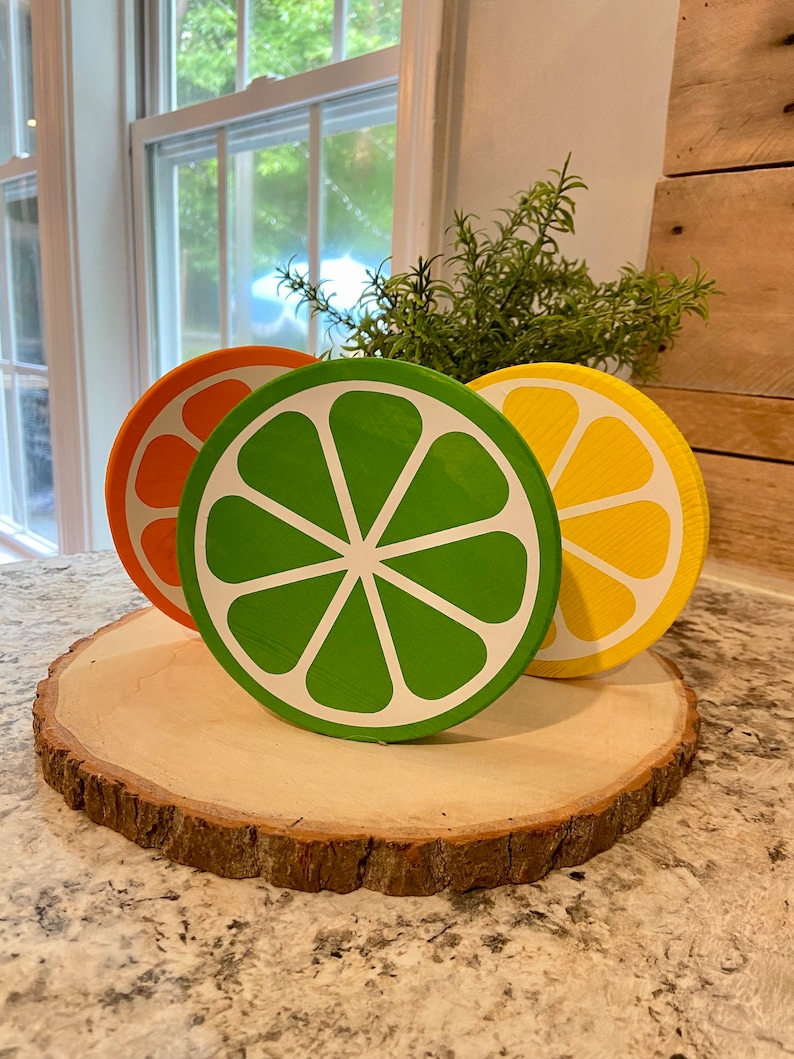 Puede incluir: Tres rodajas de madera pintadas para que parezcan frutas c&iacute;tricas. Las rodajas son naranja, verde y amarilla y est&aacute;n dispuestas sobre una rodaja de madera con corteza.