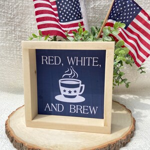 Pu&ograve; includere: Un cartello in legno incorniciato con sfondo blu e testo bianco che recita "Red, White, and Brew". Il cartello presenta una tazza di caff&egrave; bianca con stelle. Il cartello &egrave; appoggiato su una fetta di legno con due piccole bandiere americane dietro.