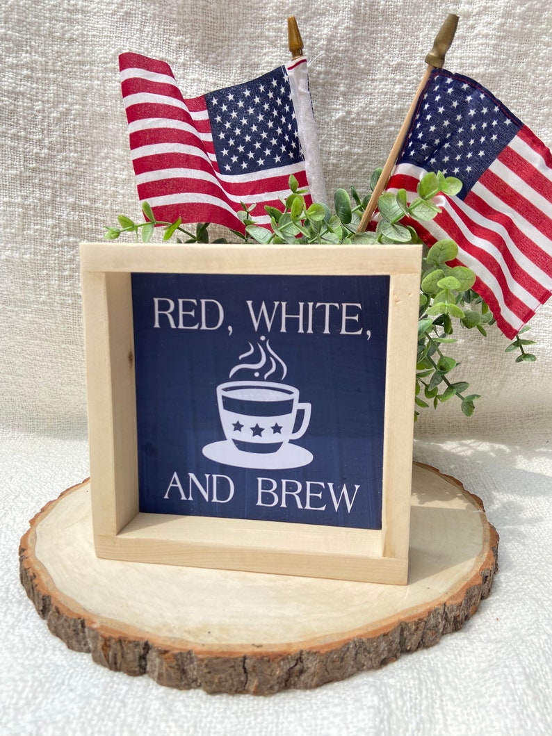 Pu&ograve; includere: Un cartello in legno incorniciato con sfondo blu e testo bianco che recita "Red, White, and Brew". Il cartello presenta una tazza di caff&egrave; bianca con stelle. Il cartello &egrave; appoggiato su una fetta di legno con due piccole bandiere americane dietro.