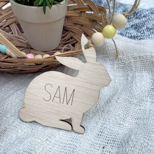 K&ouml;nnte beinhalten: Ein ausgeschnittener Holzhase mit dem eingravierten Namen "SAM". Der Hase sitzt auf einer wei&szlig;en Stoffoberfl&auml;che.