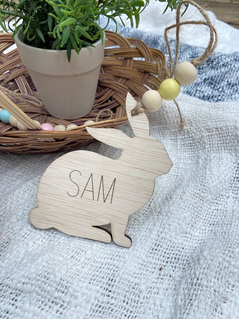 K&ouml;nnte beinhalten: Ein ausgeschnittener Holzhase mit dem eingravierten Namen "SAM". Der Hase sitzt auf einer wei&szlig;en Stoffoberfl&auml;che.