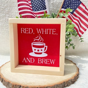 Pu&ograve; includere: Un cartello di legno con sfondo rosso e testo bianco che recita "Red, White, and Brew". Una tazza di caff&egrave; bianca con stelle e vapore &egrave; al centro del cartello. Due piccole bandiere americane sono sullo sfondo.