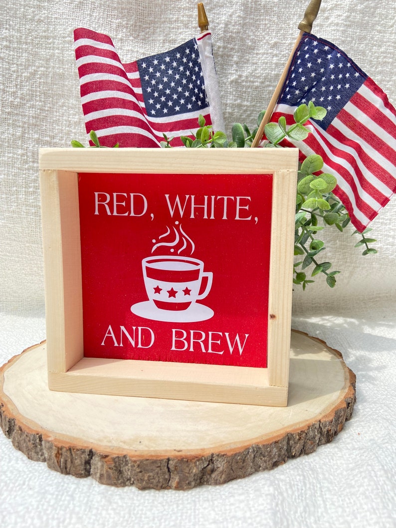 Pu&ograve; includere: Un cartello di legno con sfondo rosso e testo bianco che recita "Red, White, and Brew". Una tazza di caff&egrave; bianca con stelle e vapore &egrave; al centro del cartello. Due piccole bandiere americane sono sullo sfondo.
