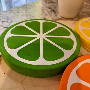 Puede incluir: Tres rodajas de fruta c&iacute;trica de madera, pintadas en verde, amarillo y naranja, con contornos blancos. Las rodajas est&aacute;n dispuestas sobre una superficie de madera.