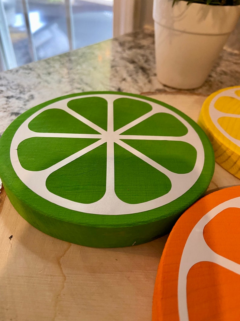 Puede incluir: Tres rodajas de fruta c&iacute;trica de madera, pintadas en verde, amarillo y naranja, con contornos blancos. Las rodajas est&aacute;n dispuestas sobre una superficie de madera.