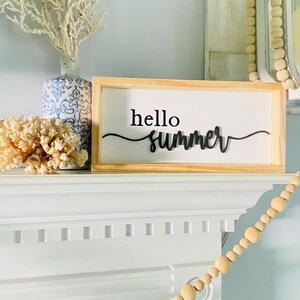Op de afbeelding: Een houten bord met de tekst "hello summer" in zwarte cursieve letters. Het bord staat op een witte schouw met een witte vaas en koraal links en een houten kralensnoer rechts.