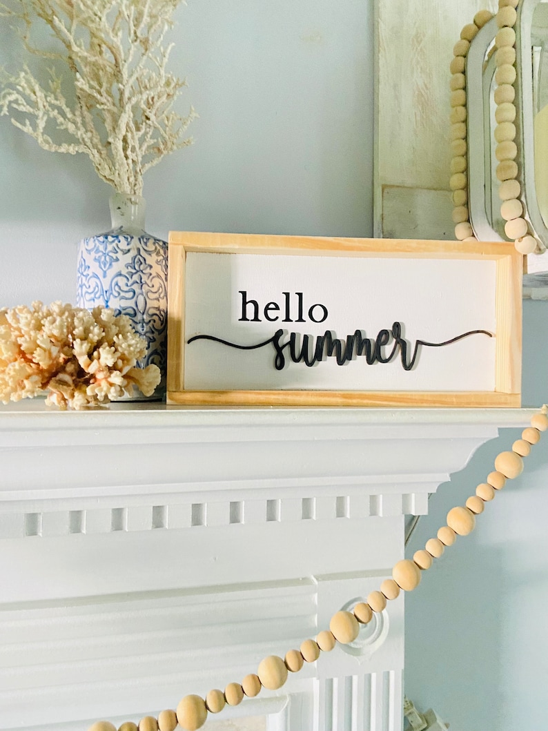 Op de afbeelding: Een houten bord met de tekst "hello summer" in zwarte cursieve letters. Het bord staat op een witte schouw met een witte vaas en koraal links en een houten kralensnoer rechts.
