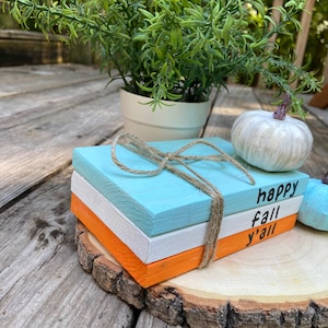 Puede incluir: Tres bloques de madera apilados sobre una rodaja de madera. Los bloques est&aacute;n pintados en tonos de azul, blanco y naranja. El bloque superior tiene el texto "happy fall y'all" escrito en negro.