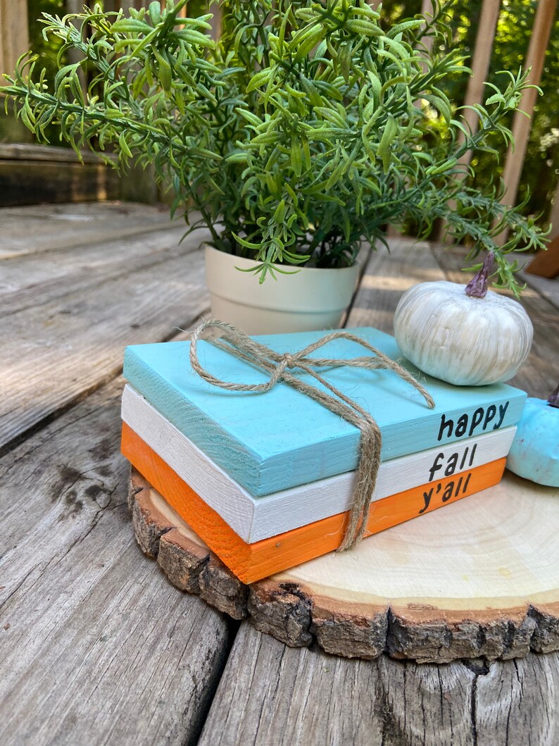 Puede incluir: Tres bloques de madera apilados sobre una rodaja de madera. Los bloques est&aacute;n pintados en tonos de azul, blanco y naranja. El bloque superior tiene el texto "happy fall y'all" escrito en negro.