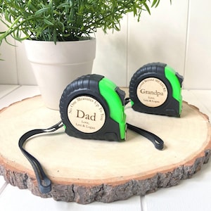 May include: Two black and green tape measures with personalized wooden labels that read "Dad" and "Grandpa". The tape measures are on a wooden slice with a small potted plant in the background. The text on the tape measures reads "No One Measures Up To You" and "Loved Beyond Measure".