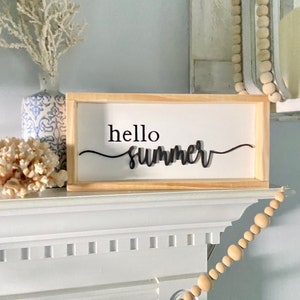 Op de afbeelding: Een houten bord met de tekst "hello summer" in zwarte cursieve letters. Het bord staat op een witte schouw met een houten kralenslinger die eraan hangt.
