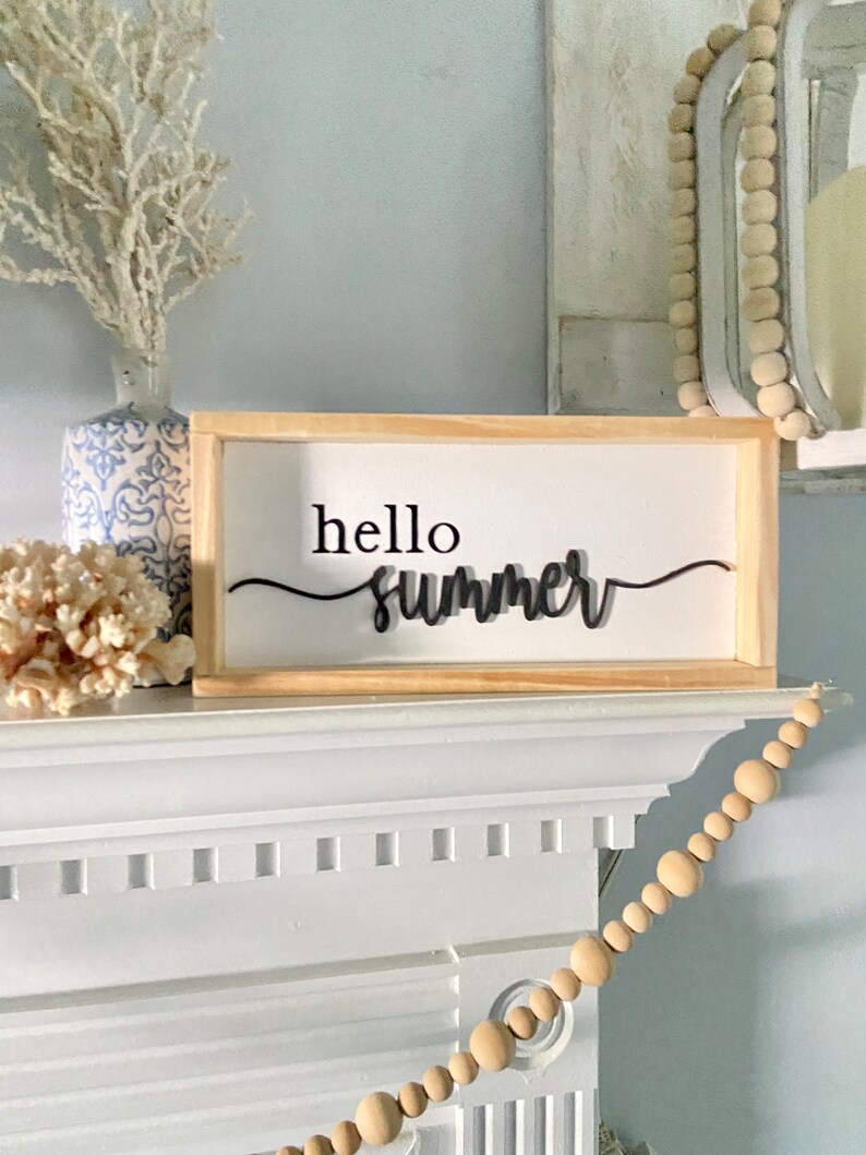 Op de afbeelding: Een houten bord met de tekst "hello summer" in zwarte cursieve letters. Het bord staat op een witte schouw met een houten kralenslinger die eraan hangt.