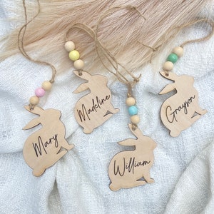 K&ouml;nnte beinhalten: Vier h&ouml;lzerne Hasen-Anh&auml;nger mit den Namen "Mary", "Madeline", "William" und "Grayson" eingraviert. Jeder Hase hat eine Schlaufe zum Aufh&auml;ngen und eine farbige Holzperle.