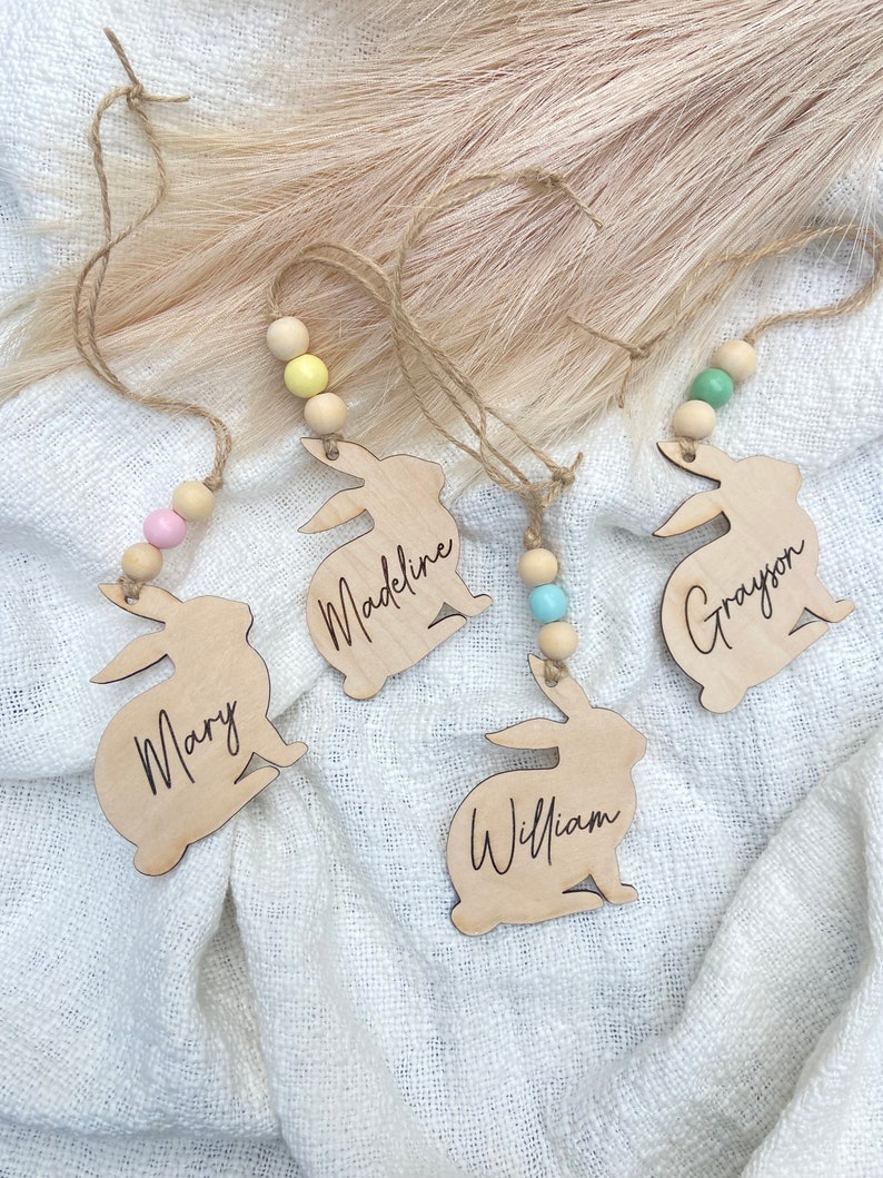 K&ouml;nnte beinhalten: Vier h&ouml;lzerne Hasen-Anh&auml;nger mit den Namen "Mary", "Madeline", "William" und "Grayson" eingraviert. Jeder Hase hat eine Schlaufe zum Aufh&auml;ngen und eine farbige Holzperle.