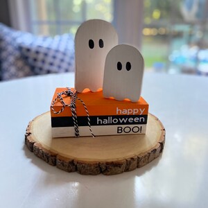 May include: A wooden ghost figurine with a smaller ghost figurine on top. The ghosts are white with black eyes. The figurines are sitting on a wooden round slice with a stack of three orange, white, and black boxes that say "happy halloween BOO!"