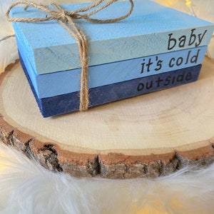 May include: Three wooden blocks stacked on top of each other, painted in shades of blue. The top block reads "baby", the middle block reads "it's cold", and the bottom block reads "outside". The blocks are tied together with twine.