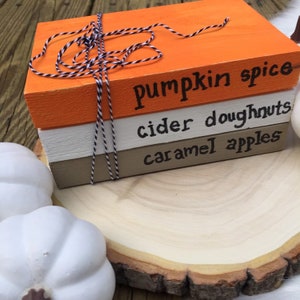 K&ouml;nnte beinhalten: Ein Stapel aus drei Holzbl&ouml;cken, die in Orange, Wei&szlig; und Braun mit schwarzer Schrift bemalt sind. Der obere Block lautet "pumpkin spice", der mittlere Block lautet "cider doughnuts" und der untere Block lautet "caramel apples".