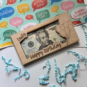 May include: A wooden money holder engraved with "Emily" and "Happy Birthday", featuring a balloon design. It holds a twenty-dollar bill. The background shows colourful wrapping paper with speech bubbles and blue paper streamers, creating a festive birthday theme.