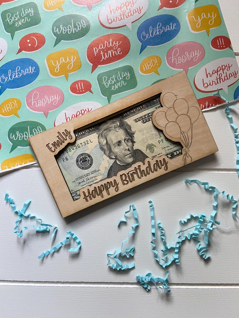May include: A wooden money holder engraved with "Emily" and "Happy Birthday", featuring a balloon design. It holds a twenty-dollar bill. The background shows colourful wrapping paper with speech bubbles and blue paper streamers, creating a festive birthday theme.