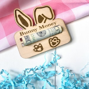 May include: A wooden "Bunny Money" holder with bunny ears and paw prints, holding a rolled-up dollar bill. The holder is on a white surface with blue paper shreds and a pink and white checkered background.