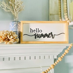 Op de afbeelding: Een houten bord met de tekst "hello summer" in zwarte letters. Het bord staat op een witte schouw met een witte koraal en een blauw-witte keramische vaas met een witte tak erachter. Een houten kralenslinger hangt aan de schouw.