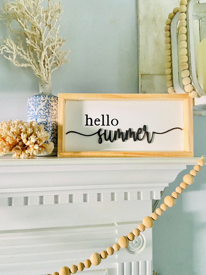 Op de afbeelding: Een houten bord met de tekst "hello summer" in zwarte letters. Het bord staat op een witte schouw met een witte koraal en een blauw-witte keramische vaas met een witte tak erachter. Een houten kralenslinger hangt aan de schouw.