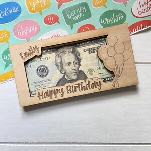 May include: A wooden money holder engraved with "Happy Birthday" and "Emily". It's designed to hold a twenty-dollar bill, with a laser-cut balloon design. The background features colourful speech bubbles with celebratory phrases.