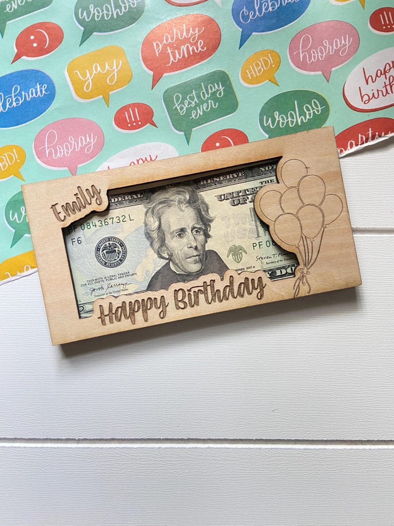 May include: A wooden money holder engraved with "Happy Birthday" and "Emily". It's designed to hold a twenty-dollar bill, with a laser-cut balloon design. The background features colourful speech bubbles with celebratory phrases.
