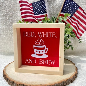 Pu&ograve; includere: Un cartello di legno con il testo "Red, White, and Brew" in lettere bianche su sfondo rosso. Una tazza di caff&egrave; bianca con tre stelle &egrave; raffigurata sopra il testo. Il cartello &egrave; appoggiato su una fetta di legno con due piccole bandiere americane dietro.
