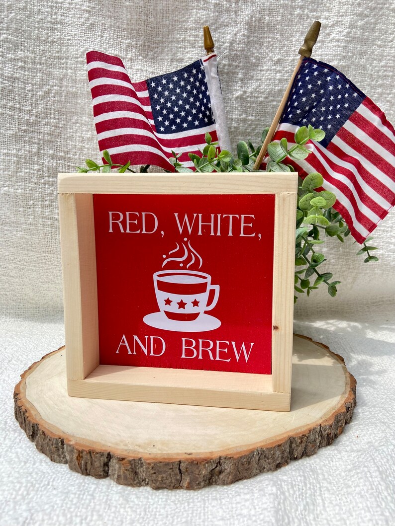 Pu&ograve; includere: Un cartello di legno con il testo "Red, White, and Brew" in lettere bianche su sfondo rosso. Una tazza di caff&egrave; bianca con tre stelle &egrave; raffigurata sopra il testo. Il cartello &egrave; appoggiato su una fetta di legno con due piccole bandiere americane dietro.